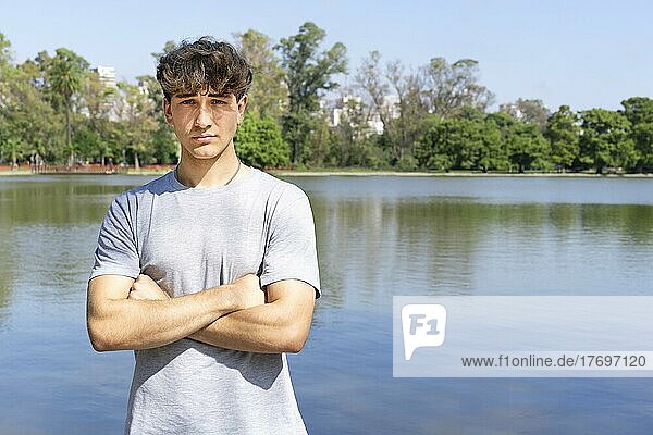 Portrait of confident young blond man with crossed arms looking at camera  summer lake background. Copy space