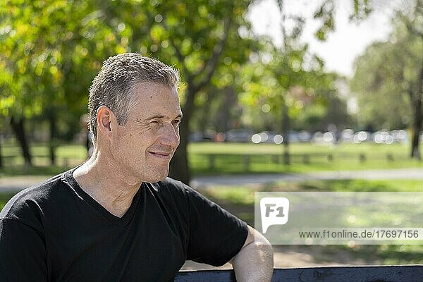 Nahaufnahme eines reifen Mannes mit grauem Haar  der ein schwarzes T-Shirt trägt und auf einer Parkbank sitzt und zur Seite schaut