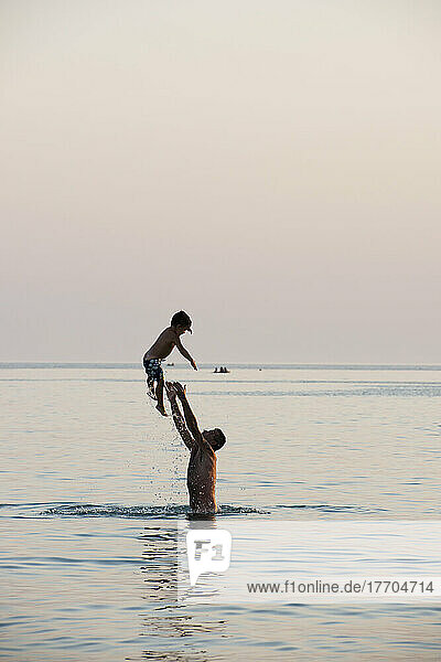 A Father Standing In The Water And Throwing His Son Into The Air; Puglia  Italy