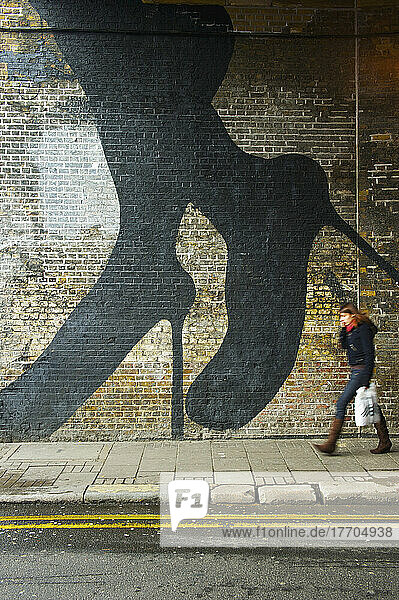 Ein Fußgänger geht an einer Backsteinmauer mit den Beinen und Füßen einer Frau in Stöckelschuhen vorbei  die auf die Wand gemalt sind  Shoreditch; London  England