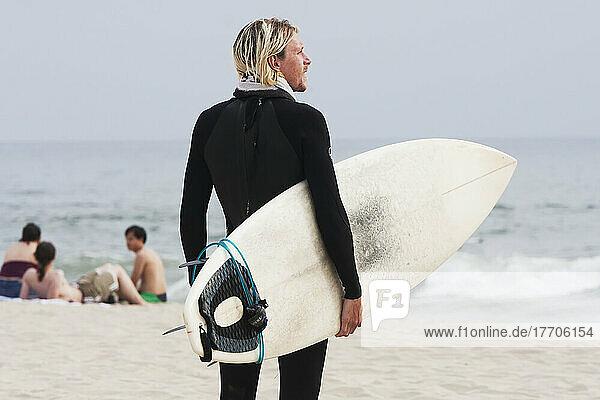 A Surfer Stands On The Beach Holding His Surfboard And Looking Out At The Water; California  United States Of America