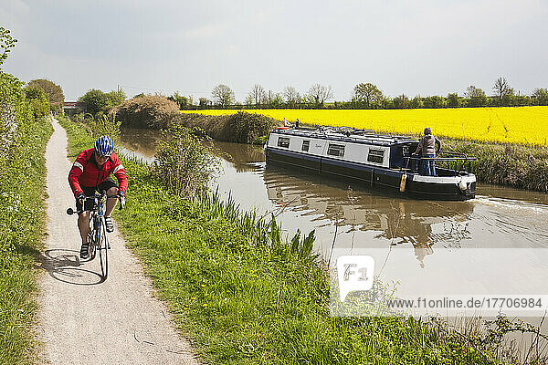 Radfahrer Radfahren entlang Kanal Seite in offenen Landschaft vorbei Narrowboat und Raps Ölsaat Felder; Wiltshire  England