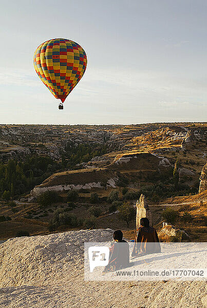 Ein farbenfroher Heißluftballon im Flug  während zwei Menschen auf einem Felsen sitzen und zusehen; Goreme  Kappadokien  Türkei