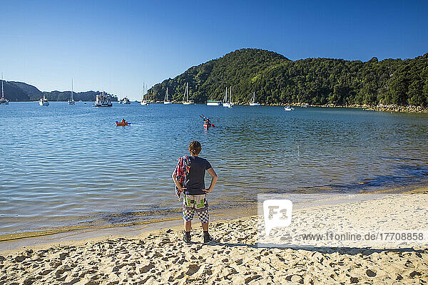Man standing on the golden sand beach watching friends in a canoe paddling back to shore on a sunny day; Abel Tasman National Park  Tasman  New Zealand