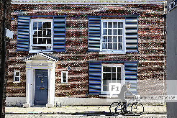 Eine junge Frau auf einem Fahrrad fährt an einem Haus mit blauen Fensterläden und blauer Tür vorbei; Winchester  Hampshire  England