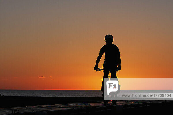 Silhouette eines jungen Mannes auf einem Stuntfahrrad im BMX-Stil  der bei Sonnenuntergang an der West Bay  an der Jurassic Coast  South West Coast Path  Dorset  England  Fahrradstunts und -tricks vorführt