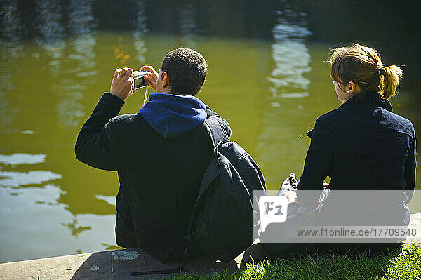 Ein junges Paar sitzt am Rande des Wassers und macht Fotos; London  England