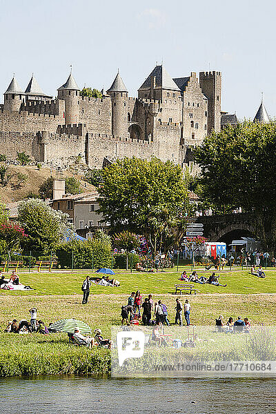 Menschen versammeln sich auf der Wiese am Fluss mit dem Schloss und den Festungsmauern im Hintergrund; Carcassonne  Languedoc-Rousillion  Frankreich