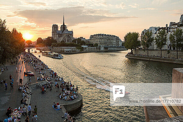 The sun sets near the Notre Dame Cathedral as boats cruise along the Seine and Parisians sit along the river banks.; Paris  France