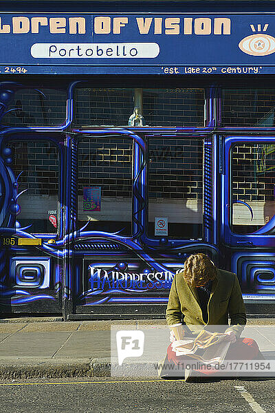 Ein Mann sitzt und liest eine Zeitung am Straßenrand  Portobello Road Market; London  England
