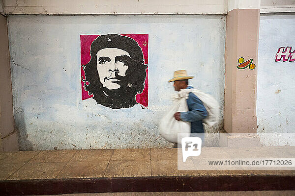 Ein Mann trägt eine Last neben einem Bild von Che Guevara an der Wand eines kubanischen Bauernmarktes; Cienfuegos  Kuba