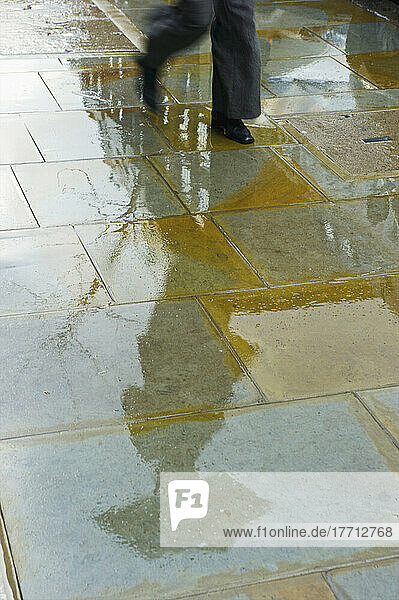 Ein Fußgänger geht mit einem Regenschirm auf dem nassen Gehweg mit ihrer Reflexion in einer Pfütze; London  England
