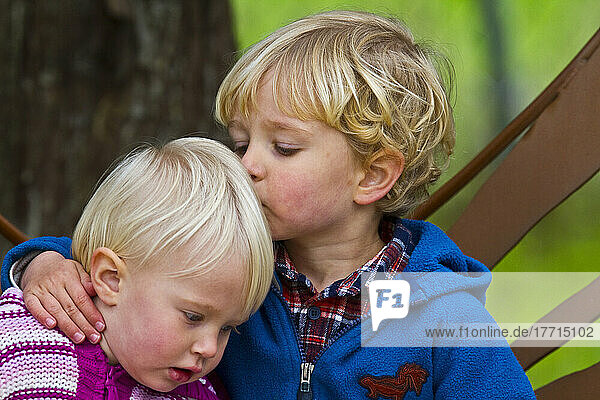 Brother Kissing Sister On The Head Sitting On A Bench.