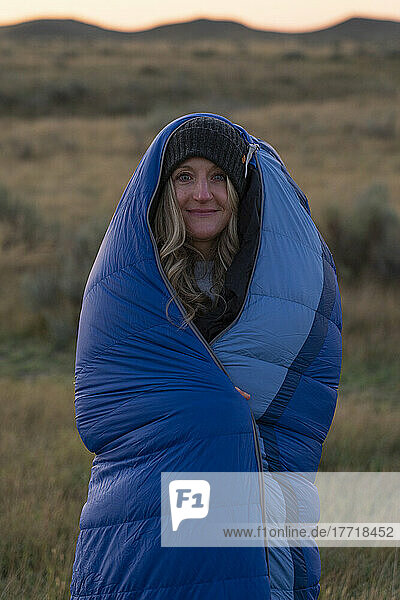 Eine Frau wickelt sich bei Sonnenaufgang in einen Schlafsack ein  um sich warm zu halten  während sie in der weiten Prärielandschaft steht; Val Marie  Saskatchewan  Kanada