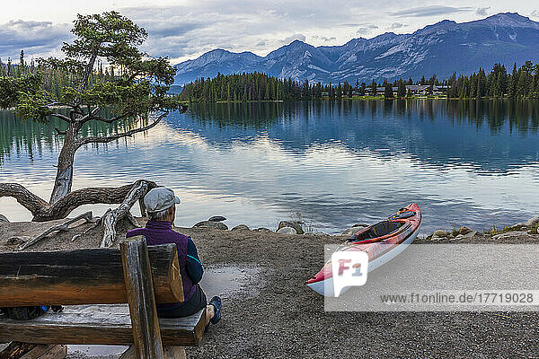 Woman on bench and kayak on shore of Beauvert Lake in Jasper National Park with the Fairmont Jasper Park Lodge in the background; Alberta  Canada
