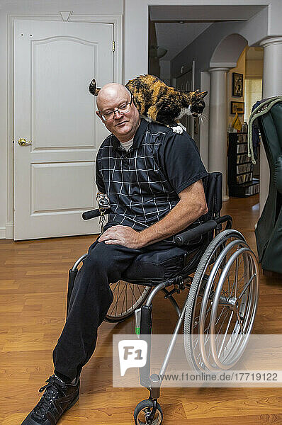 Mann mit doppelter Gliedmaßenamputation  der in seinem Haus sitzt  während seine Hauskatze über seine Schultern krabbelt; St. Albert  Alberta  Kanada