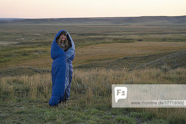 Woman wrapped up in a sleeping bag at sunrise to keep warm as she stands on the vast prairie landscape; Val Marie  Saskatchewan  Canada