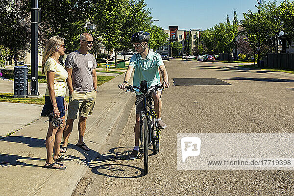 Junger Mann  der mit seinem Fahrrad eine Wohnstraße hinunterfährt und anhält  um mit seinen Eltern zu sprechen  die auf dem Bürgersteig stehen; Edmonton  Alberta  Kanada