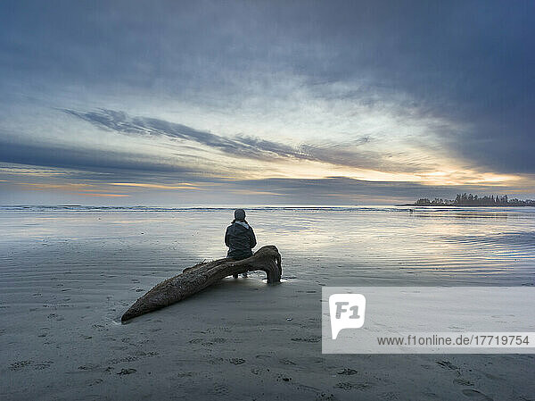 A woman sits on driftwood and enjoys watching the sunset on Long Beach  the largest and longest beach in the Pacific Rim National Park Reserve on the west coast of Vancouver Island; British Columbia  Canada