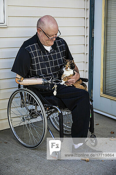 Mann mit doppelter Gliedmaßenamputation sitzt vor seinem Haus mit seiner Hauskatze auf dem Schoß; St. Albert  Alberta  Kanada