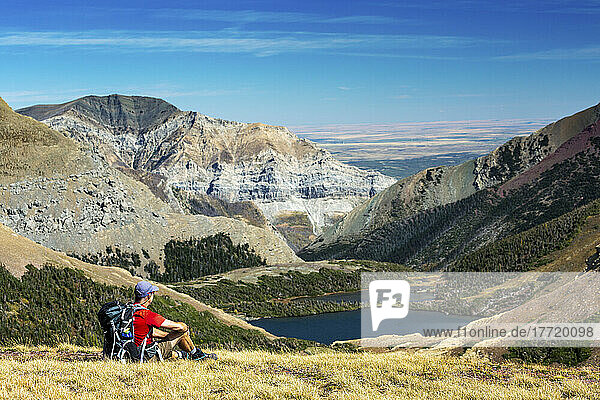 Männlicher Wanderer  der sich auf einem grasbewachsenen Bergrücken ausruht  mit Blick auf einen Alpensee  Bergketten  blauer Himmel und Wolken im Hintergrund  Waterton Lakes National Park; Waterton  Alberta  Kanada
