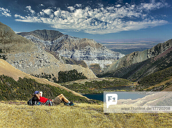 Male hiker relaxing on grassy mountain ridge overlooking an alpine lake  mountain ranges  blue sky and clouds in the background  Waterton Lakes National Park; Alberta  Canada
