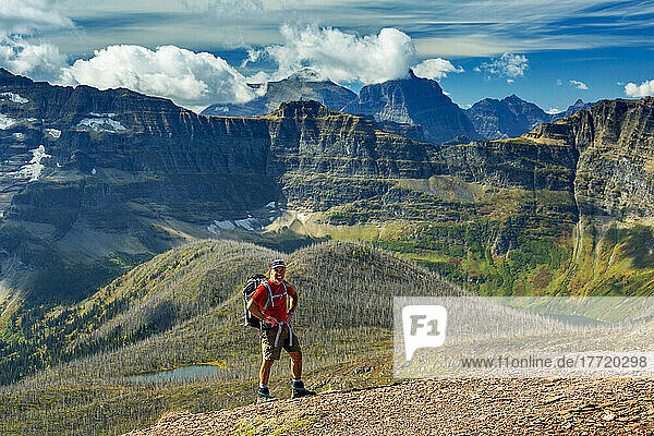 Male hiker on top of a mountain ridge overlooking a valley below and mountain ranges in the background with blue sky and clouds  Waterton Lakes National Park; Alberta  Canada