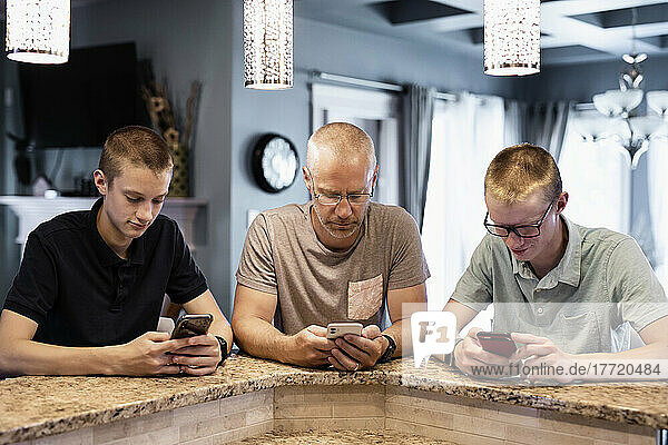 Vater und zwei Söhne sitzen an der Kücheninsel zu Hause und benutzen ihre Smartphones; Edmonton  Alberta  Kanada