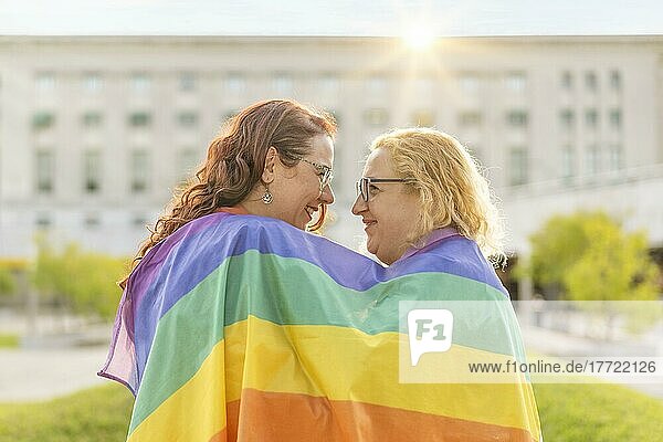 Verliebtes lesbisches Frauenpaar  das sich gegenseitig umarmt  eingewickelt in eine schwule Flagge  in einem Park bei Sonnenuntergang  mit Reflexionen der Sonne. Konzept der Vielfalt  Stolz  Liebe  Gleichheit