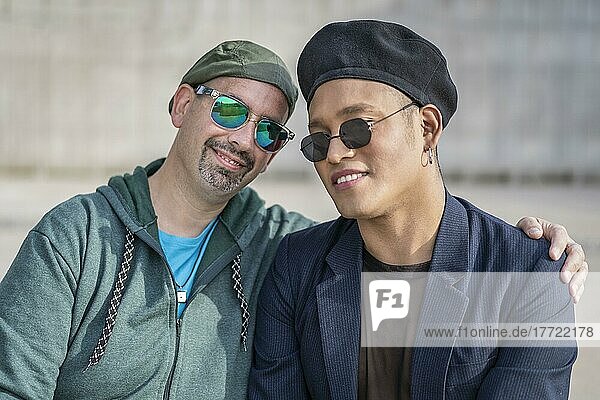 Gay Latino male couple sitting on a bench in a park  wearing fashionable hats and sunglasses  looking at the camera smiling