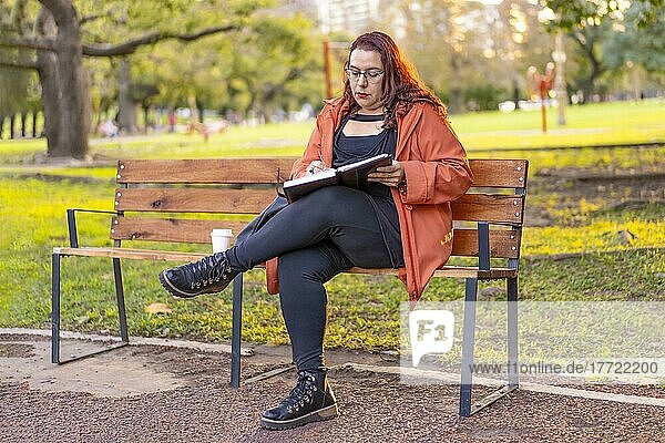 Frau auf einer Parkbank sitzend  die ihren Terminkalender und ihr Handy überprüft  während sie einen Kaffee trinkt