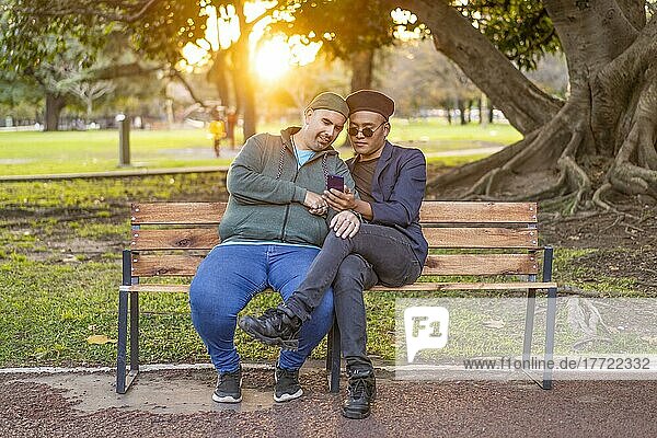 Homosexuelles lateinamerikanisches Paar  das bei Sonnenuntergang auf einer Bank in einem Park sitzt  modische Hüte trägt  ein Handy in der Hand hält und lächelt