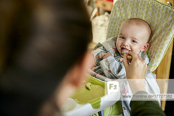 Smiling six month old baby being fed by mother in high chair
