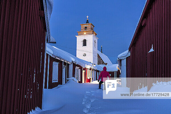Person admiring the bell tower standing in deep snow among red cottages at dusk  Gammelstad Church Town  UNESCO World Heritage Site  Lulea  Sweden  Scandinavia  Europe