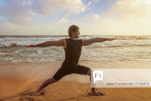 Man doing Hatha yoga asana Virabhadrasana 1 Warrior Pose outdoors on ocean beach on sunset. Kerala  India  Asia