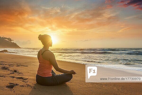 Frau macht Yoga  meditieren und entspannen in Padmasana Lotus Asana Pose mit Kinn Mudra im Freien am tropischen Strand bei Sonnenuntergang mit dramatischen Sonne