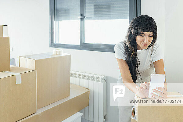 Smiling woman using mobile phone leaning on cardboard boxes at new home