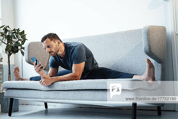 Man using smart phone sitting with legs apart on sofa at home