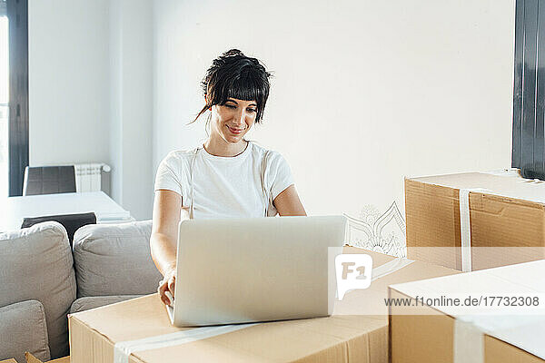 Smiling woman using laptop on cardboard box in living room at home