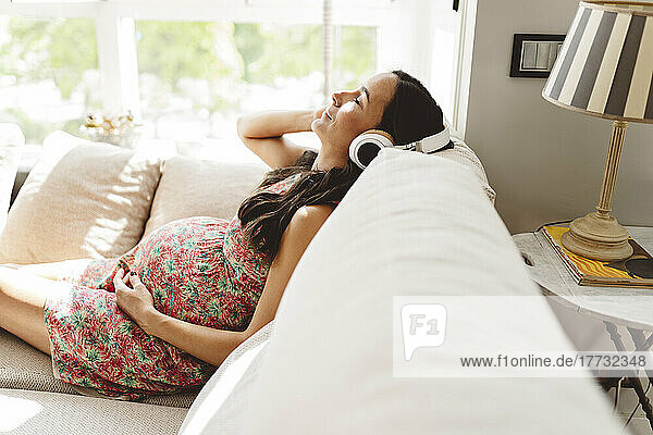 Schwangere Frau mit geschlossenen Augen hört Musik über kabellose Kopfhörer und sitzt zu Hause auf dem Sofa