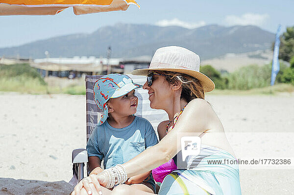 Smiling woman wearing hat sitting with son at beach on sunny day