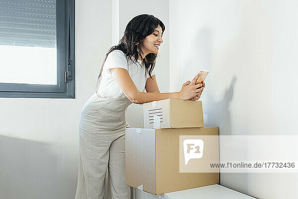 Smiling woman text messaging through smart phone leaning on cardboard boxes at home