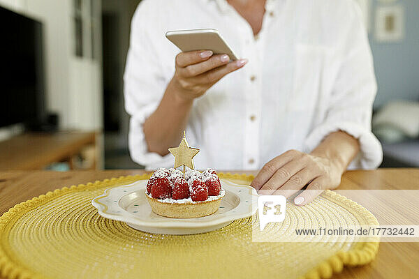 Woman photographing raspberry tart with star shape candle through smart phone at home