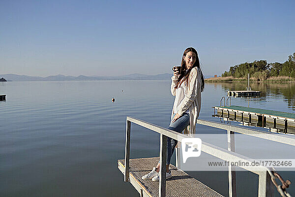 Frau mit Kaffeetasse steht auf Steg über See