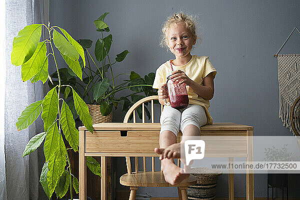 Smiling girl with jar of smoothie sitting on table at home