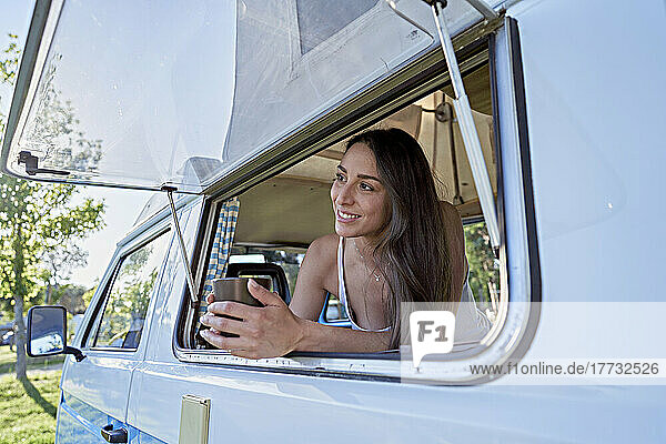 Lächelnde Frau mit Kaffeetasse  die aus dem Fenster ihres Wohnmobils schaut