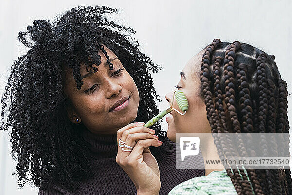 Smiling woman doing facial massage on friend through jade roller against white background