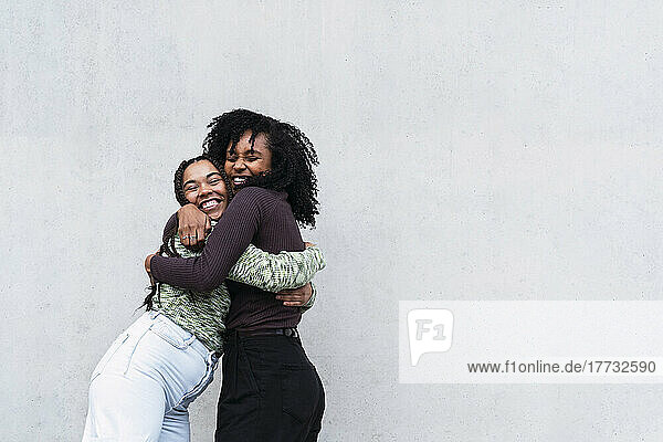 Cheerful female friends hugging each other in front of wall