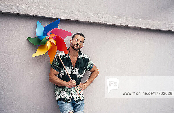 Thoughtful man with hand on hip holding pinwheel toy leaning on concrete wall