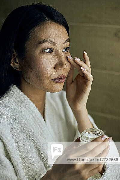 Woman wearing bathrobe applying cream on face